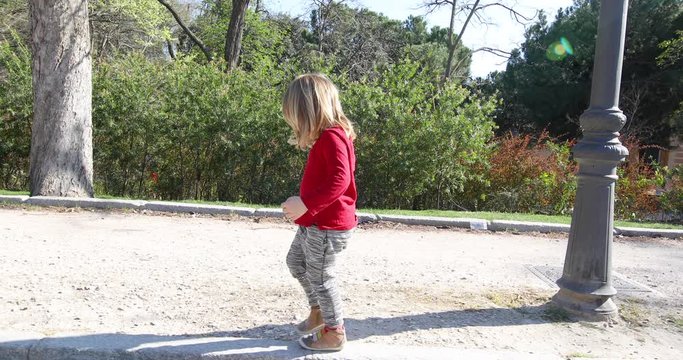 slow motion. Side view of three years old blonde child, with red shirt and grey trousers, walking balancing on curb of sidewalk in public park
