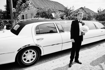 Stylish groom with wedding bouquet at hands near limousine.