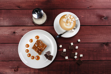 a piece of chocolate brownie and caramel sauce, a cup of cappuccino on a white plate. Top view