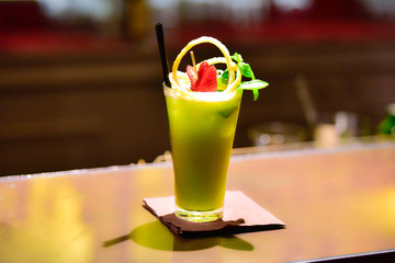 A green cocktail on the bar counter. It is decorated with fruits ( strawberry, lemon peel, mint ). Blurred background