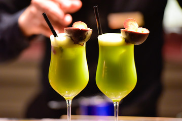 Two green cocktails on the bar counter. They are decorated with fruits and the shell of a fruit ). Blurred background with the barman