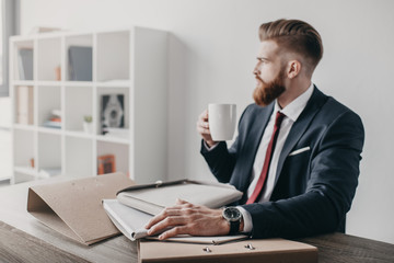 young businessman with documents and folders drinking coffee and sitting at table in office