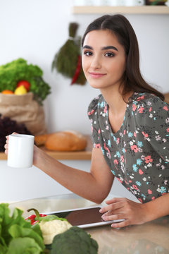 Young latin  woman is making online shopping by tablet computer while holding white cup of tee. Housewife found new recipe for  cooking in a kitchen