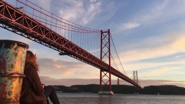 Lisbon Landmarks, Girl Sits By River Tagus During Sunset. Young woman enjoys beautiful sunset by Tagus river and famous 25 de Abril bridge in Lisbon