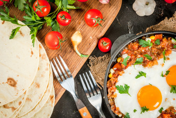 Traditional Mexican dish Huevos rancheros - scrambled eggs with tomato salsa, with taco tortillas, fresh vegetables and parsley. Breakfast for two. On a black concrete table.