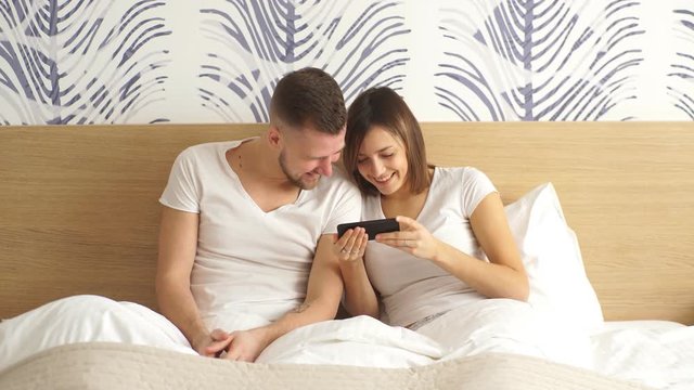 Beautiful young happy couple in bed with mobile phone smiling. Domestic atmosphere, lifestyle photo.
