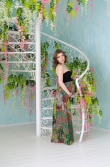 Young pregnant woman stands near a white vintage spiral staircase decorated with flowers