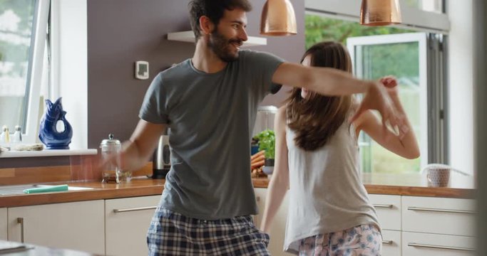 Happy young couple dancing in kitchen wearing pajamas listening to music morning at home having fun