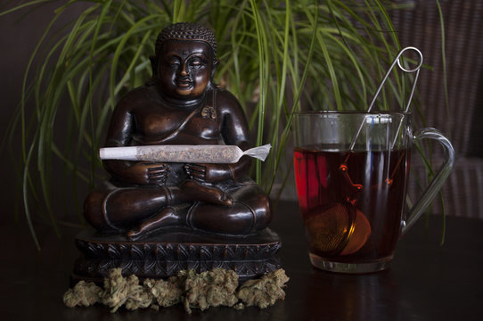 closeup of rolled marijuana weed joint and buds with Buddha statuette and cup of rooibos tea on wooden ground