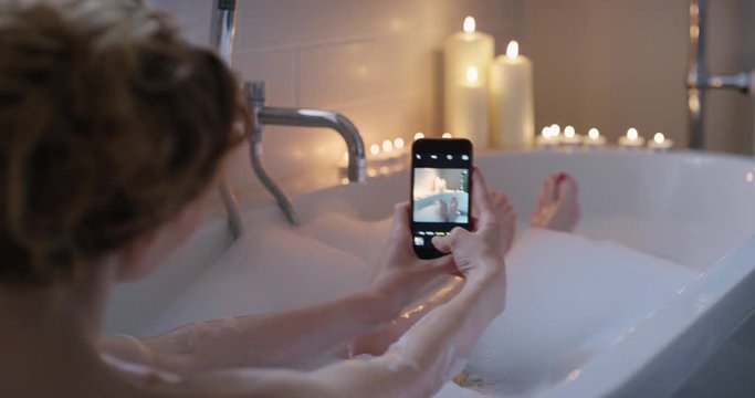 Woman lying in bubble bath photographing feet using smartphone sharing photos on social media relaxing at home mindfulness concept