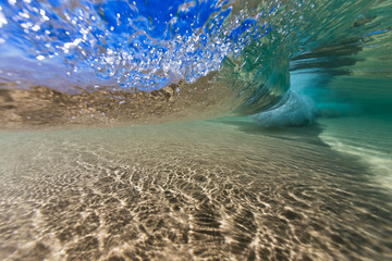 An underwater shot of beneath a wave breaking in crystal clear water over a sand bottom.