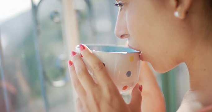 Beautiful young Woman drinking coffee alone at home looking out of window relaxing enjoying hot drink in the morning holding multi colored polka dot mug