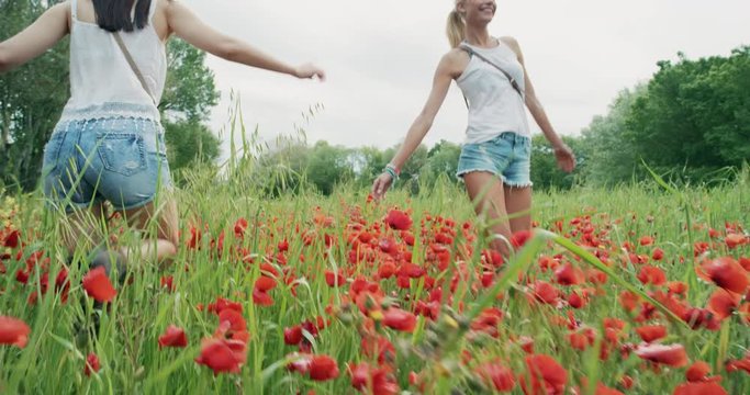 Two young girls wearing denim shorts running through field of wild poppies dancing and spinning in slow motion enjoying european travel adventure