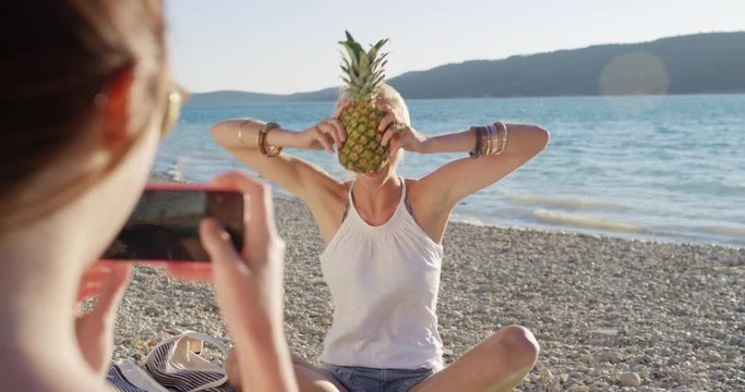 Close up attractive woman balancing pineapple on head posing for camera smiling laughing at sunset on tropical beach