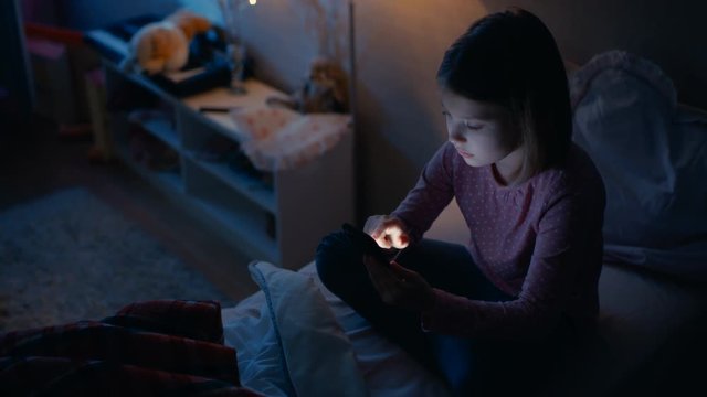 Cute Little Girl in Her Room at Night. She's Sitting on Her Bed While Using Smartphone. Her Bed Light is On. Shot on RED EPIC-W 8K Helium Cinema Camera.