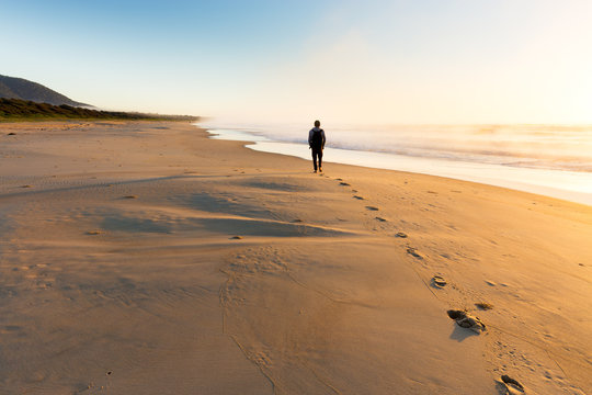 A person walking on a beautiful beach leaves a trail of footprints as the sunrise illuminates mist rising from the sea in golden light.