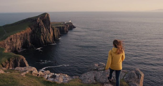 Woman with arms raised on top of cliff looking at Ocean view at sunset Hiker Girl wearing arm up celebrating landscape enjoying vacation travel adventure Neist Point Lighthouse Isle of Skye Scotland