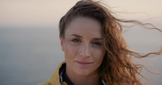 Close up portrait of Young Woman smiling with Red hair blowing in wind looking at sunset over ocean Girl wearing yellow raincoat trekking in Scotland Slow Motion