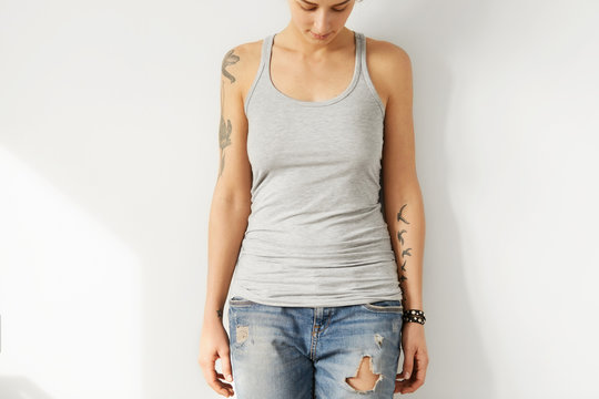 Studio shot of tattooed young European female model looking down at grey blank A-shirt. Cropped portrait of teenge girl dressed in ripped jeans and tank top posing indoors. Youth and fashion