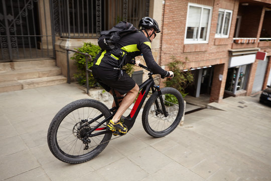 Side shot of male cyclist wearing cycling clothing and protective gear speeding on streets riding his pedal-assist electric bike, about to jump down concrete stairs. Exteme sports and adrenaline