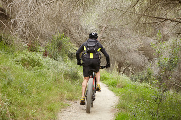 Rear view of unrecognizable young man wearing protective helmet, sports clothes and backpack speeding along forest trail on his two-wheeled motor-powered booster vehicle with pedal-assist system