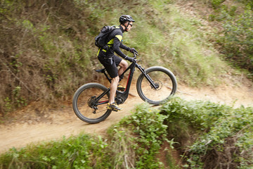 Fototapeta na wymiar Young Caucasian professional rider performing mountain biking stunts on two-wheeled motor-powered bicycle on trail along cliff using pedal-assist system. Male biker cycling outdoors on electric bike