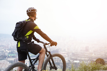 Fototapeta premium Rear shot of young rider standing on top of hill with booster bike and looking down at foggy city while biking in the morning. Cyclist in black and yellow sports clothes admiring beautiful view