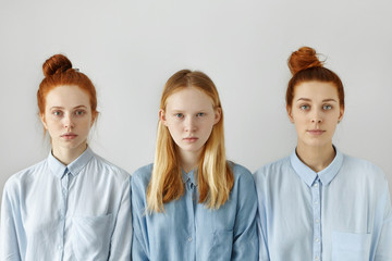 Studio shot of three college girls or sisters dressed in shirts posing against white wall background having neutral face expressions. Blond girl standing between two red haired female friends