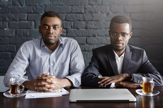 Two handsome successful Afro-American businessmen working in office sitting at table with laptop, papers and mugs, looking at camera with confident expressions against black brick wall background