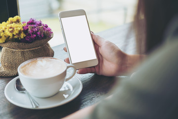 Mockup image of hands holding white mobile phone with blank white screen with hot coffee cup and flower vase in cafe
