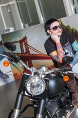 Fototapeta na wymiar Cool sensual biker girl sitting on old fashioned motorcycle in garage interior on grey wooden wall background, vertical picture