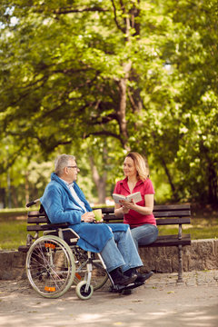 Young carer woman reading book in the park disabled man in wheelchair.