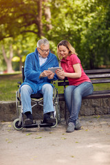 Disabled retiree man in park using tablet with daughter.