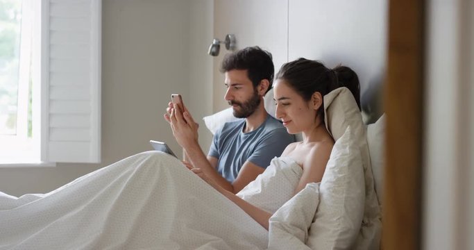 Young couple lying in bed using smartphone digital tablet technology scrolling online browsing sharing real moment morning