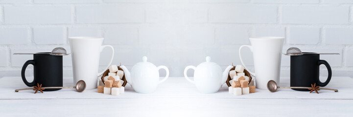 White and black coffee and tea mugs with sugar cubes and teapot on wooden table. White background. Copy space. Wide panoramic image. Horizontal banner.