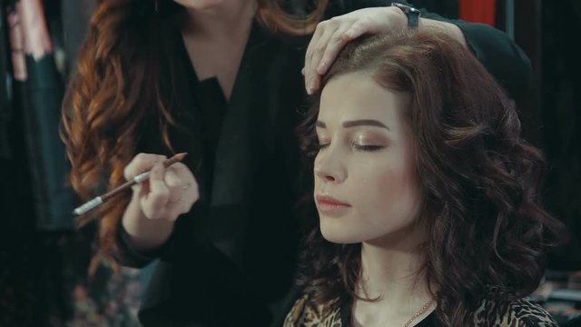 A young woman stylist puts on the eyelids of a brunette model with an eyeliner, talking about the intricacies of evening make-up.