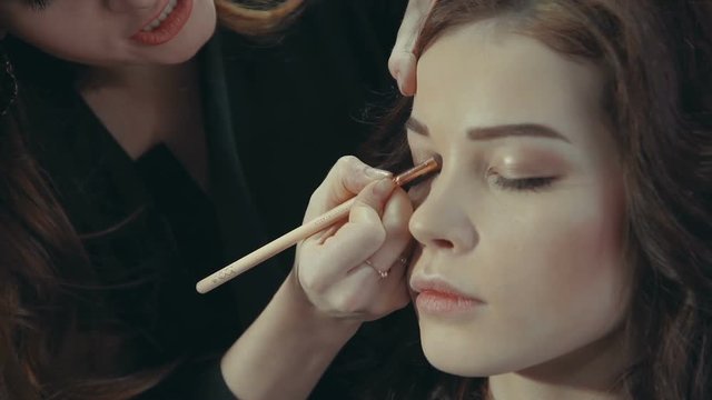 A young woman stylist puts on the eyelids of a brunette model a dark eye shadow with a special brush, talking about the intricacies of evening makeup.