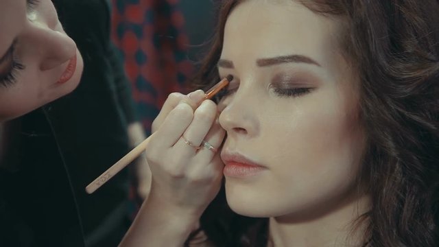A young girl stylist puts on the eyelids of a brunette model a dark eye shadow with a special brush, talking about the intricacies of evening makeup shoot close-up.