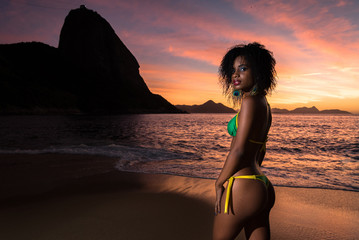 Fototapeta premium Sexy Young Brazilian Woman in Bikini Standing in the Beach by Sunrise With the Sugarloaf Mountain in the Background