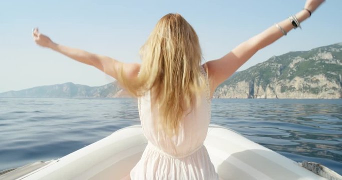 Rear view gorgeous blonde woman arms raised riding on speed boat on tropical island holiday vacation