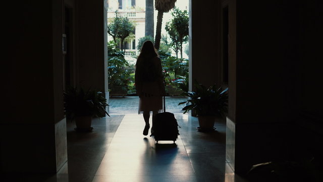 Young traveler woman walking with a suitcase in the street. Girl opens the door and comes into an arch.