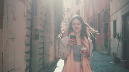 Young happy woman walking in city centre at morning, using smartphone. Girl surfing the Internet, texting with someone.