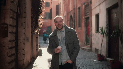 Fototapeta na wymiar Portrait of young handsome man looking at camera. Happy smiling male standing in morning street, drinking coffee.