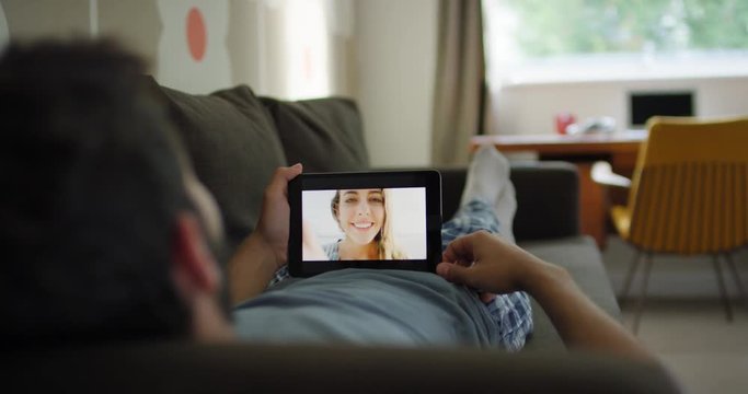 Young Man having video chat holding smartphone  webcam chatting to attractive girlfriend lying on sofa relaxing at home in modern apartment