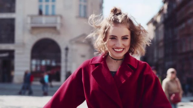 Close up view of a young playful woman walking down the street, turning to camera and laughing happily. Stylist outfit, red coat, choker, short haircut, red lips. Gorgeous woman. Happy life.