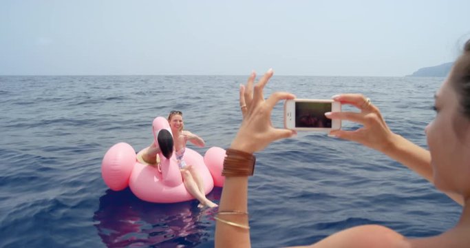 Woman lying on pink inflatable flamingo bet friend taking photograph with smartphone digital camera floating alone in middle of ocean sharing images with mobile phone technology