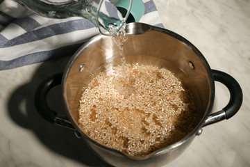 Pouring water into saucepan with brown rice on kitchen table