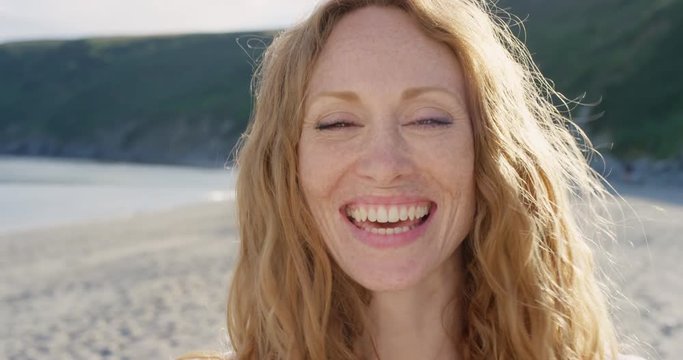 Close up portrait of beautiful young redhead woman running hand through healthy red hair blowing in wind on tropical beach slow motion