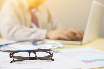 glasses on document , male doctor write prescription,medical record and computer on the table.office table desk in hospital,and other things on wooden desk background.selective focus