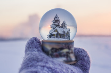 Girl in knitted mittens holding glass ball with firtrees, house and artificial snow with winter...